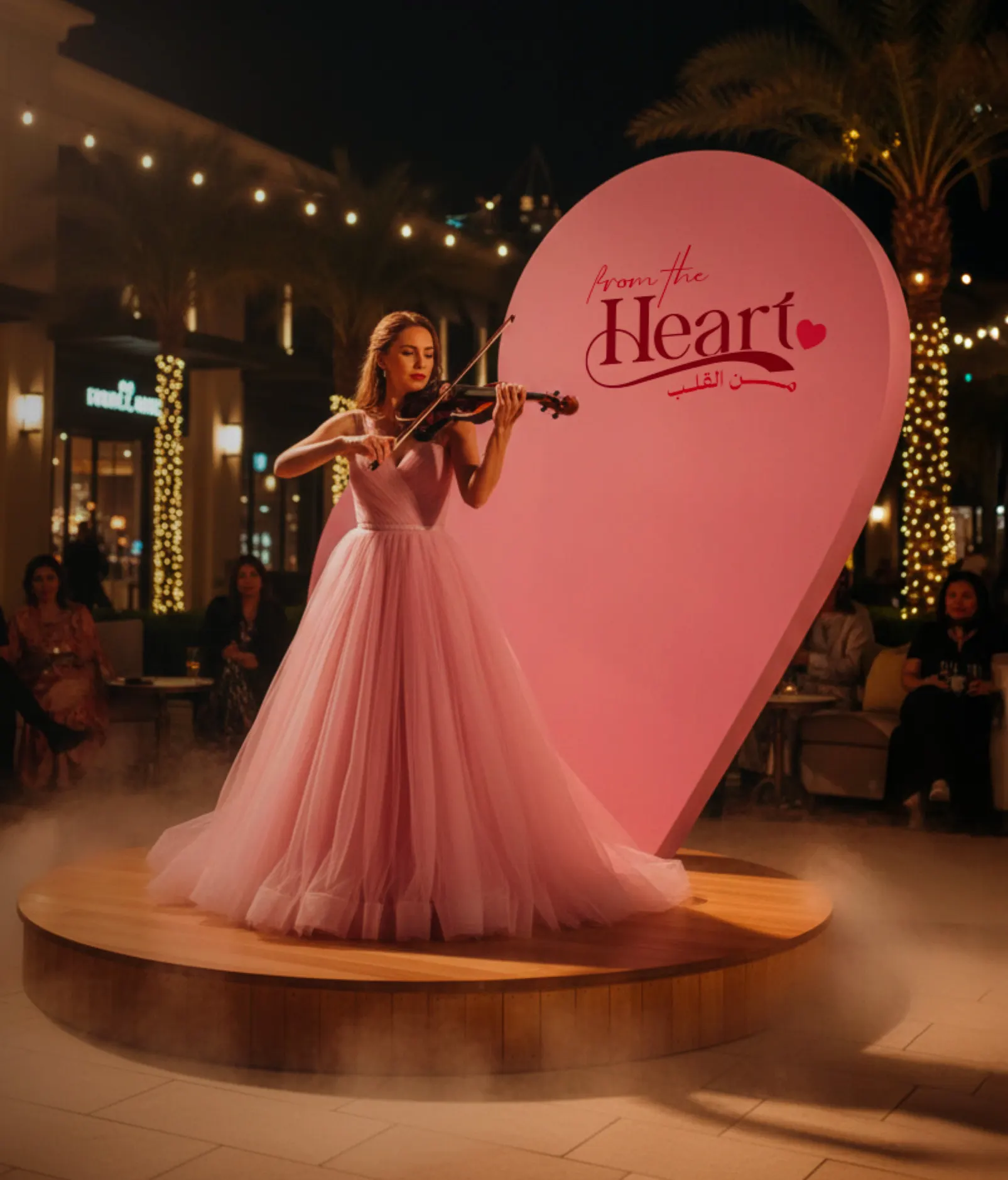 A violinist in a pink gown on a stage before a heart shaped backdrop